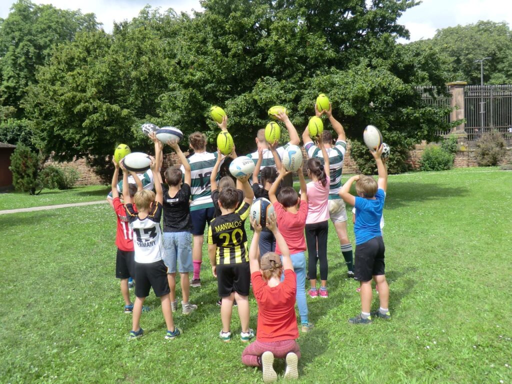 Kinder und Trainer:innen halten Rugbybälle in die Höhe.