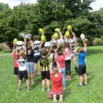 Kinder und Trainer:innen halten Rugbybälle in die Höhe.